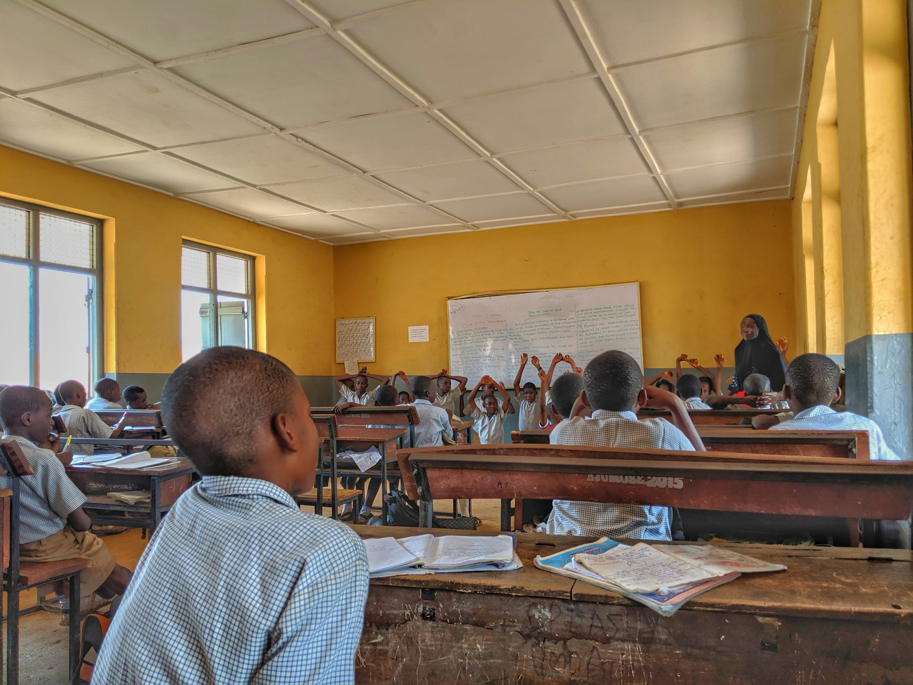 A full primary school classroom, children at wooden desks with their hands raised as a teacher takes a basic science lesson at the whiteboard.